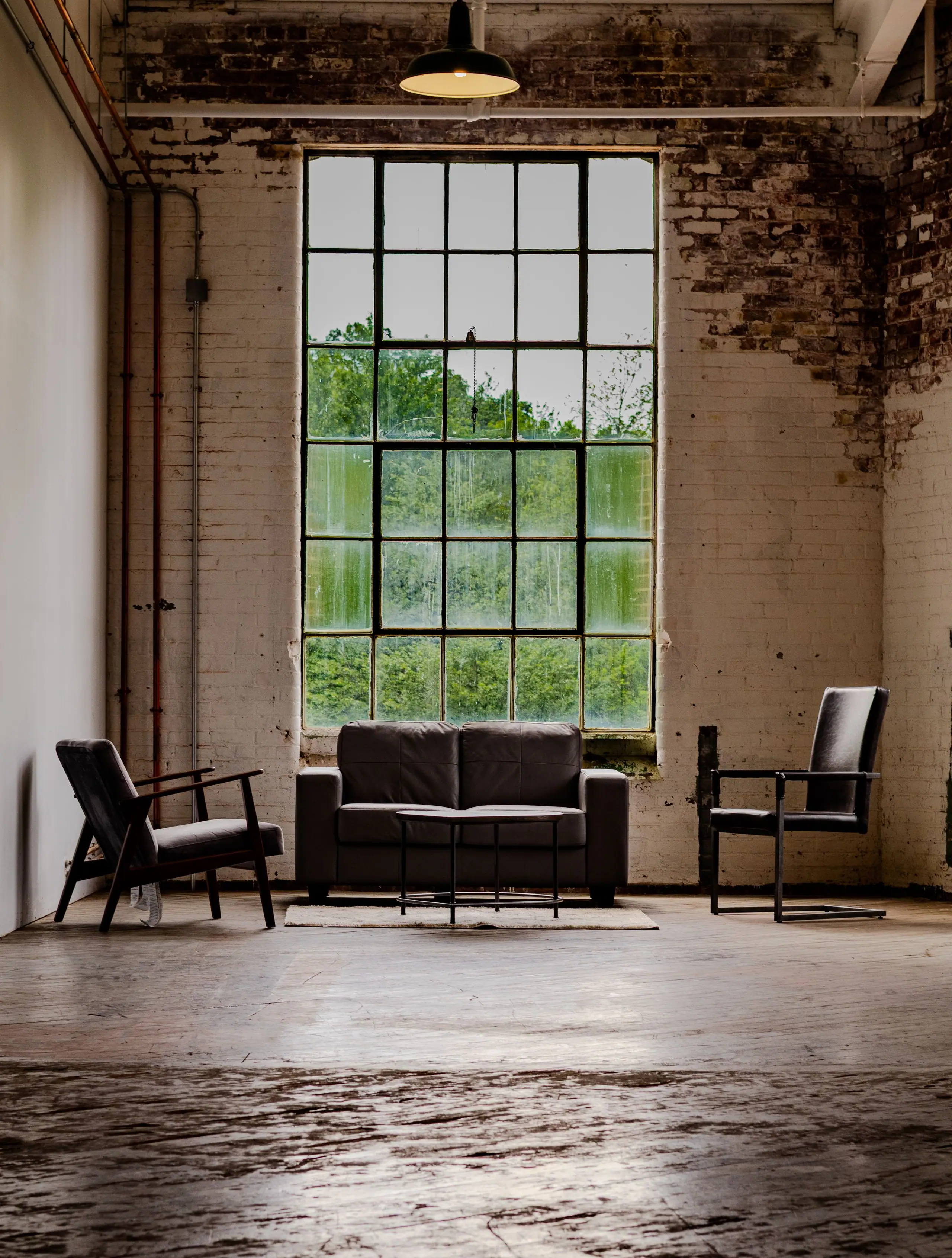 A tall cast-iron mill window with low leather chairs set on a weathered concrete floor and exposed brick walls