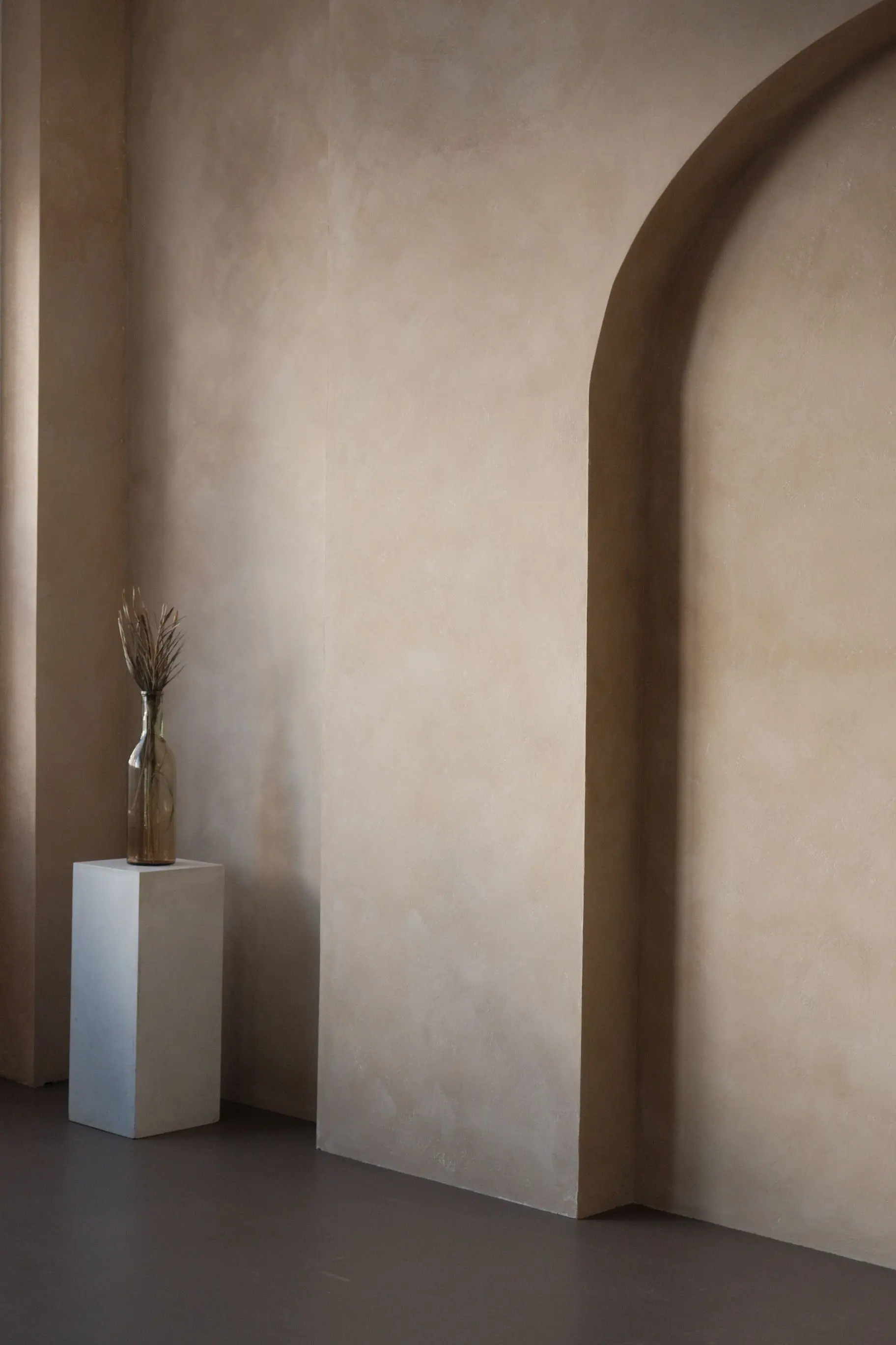 A softly lit plaster wall with a deep arched opening and a single dried stem in a glass vase