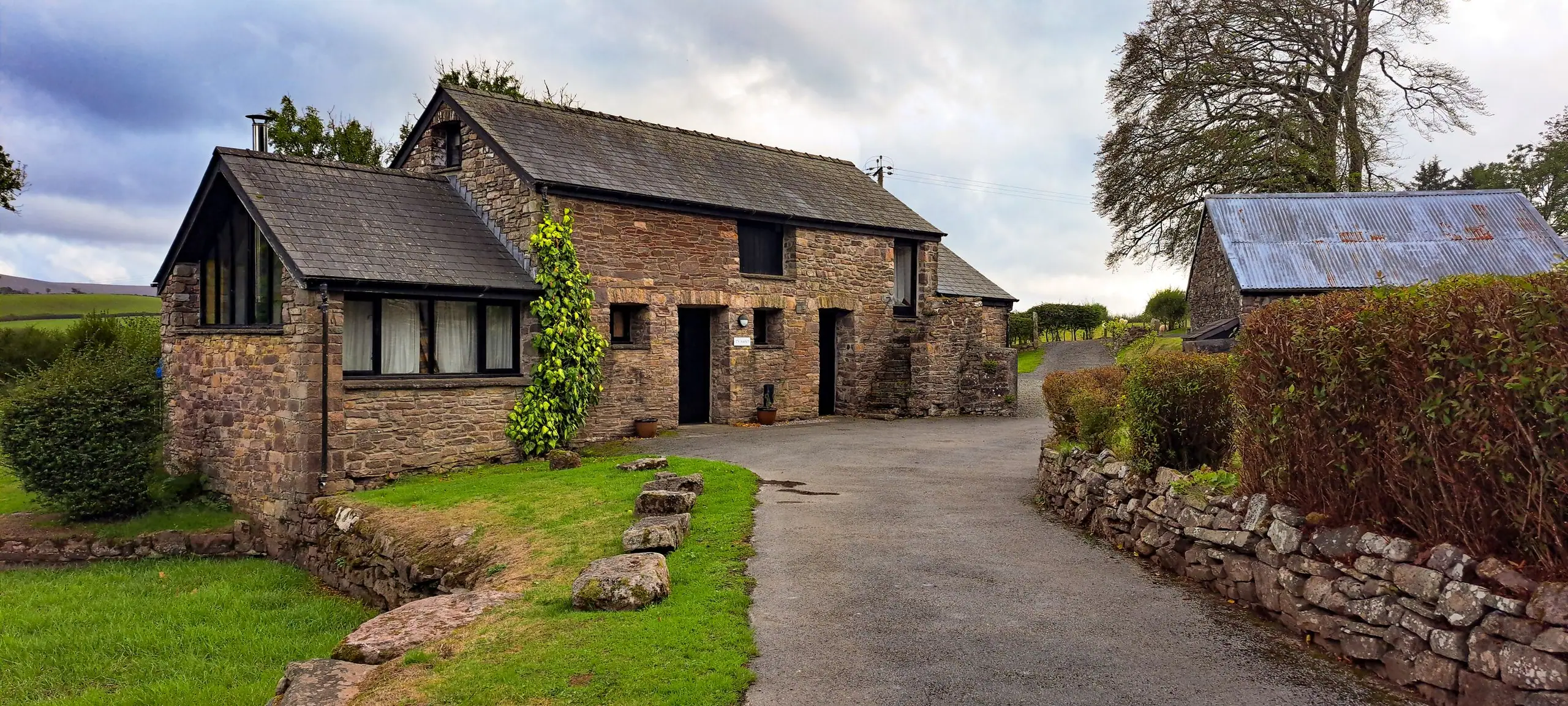 The converted gritstone barn with a new glazed gable, slate roofs and dry-stone walls along the lane
