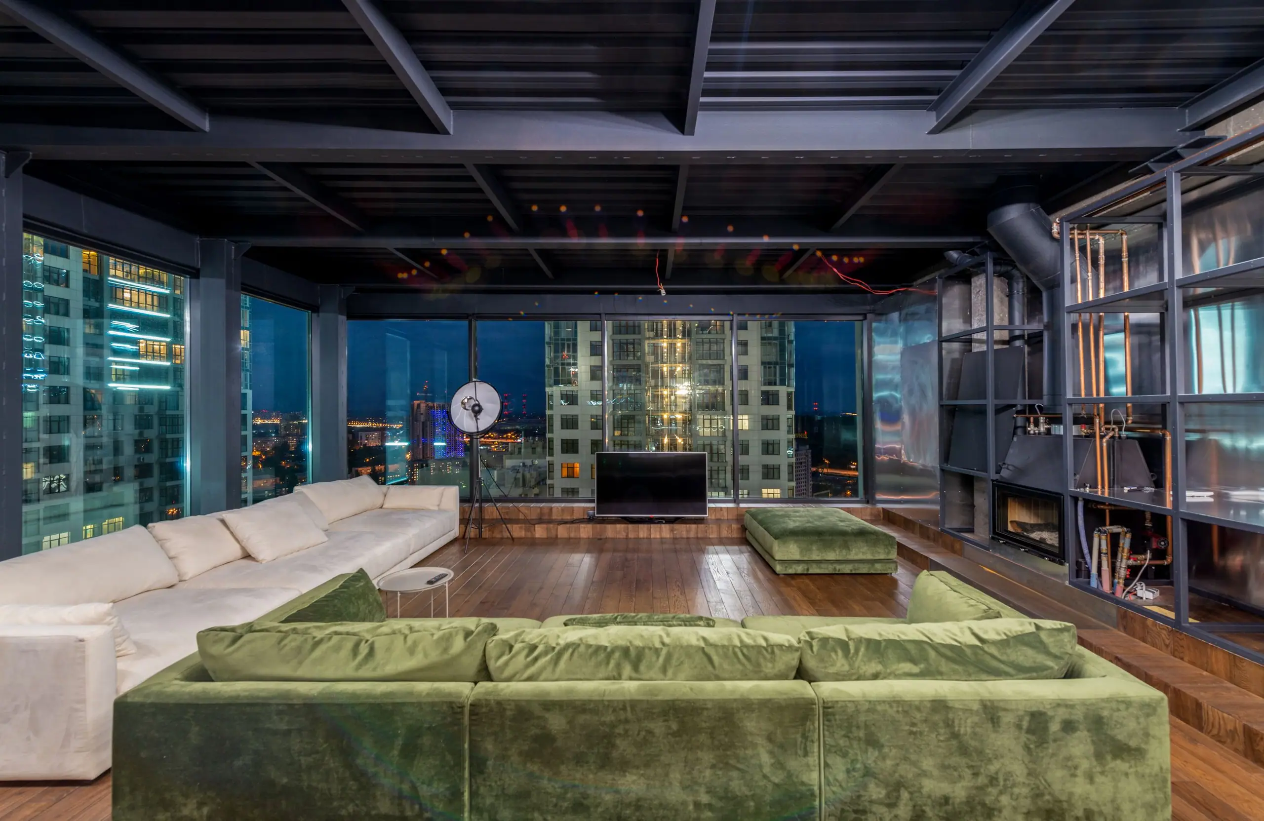 A penthouse living room at dusk with a long green velvet sofa and full-height glazing onto the lit city